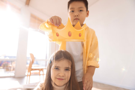 Cute children of different races play together. A small Asian boy holds a paper crown over the head of a Caucasian girl. Kids are having fun in the living room of the house.の写真素材