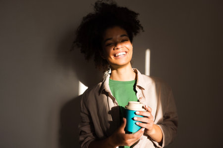 An attractive young girl of mixed race holds a cup of coffee and smiles. A charming multiethnic teenager in casual clothes is drinking tea to take away. Beautiful African woman with a hot drink.の写真素材