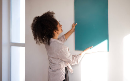 A young beautiful mixed-race girl in casual clothes applies a painted sample to the wall. A cute African woman chooses a paint color. An attractive multiethnic girl makes repairs in the house.の写真素材