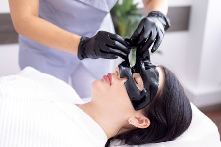 Beautiful caucasian woman with black silicone mask for the skin. Pretty brunette female with cosmetic mask on her face. Skin care cosmetics on background. Dark blackdrop. Copy spaceの写真素材