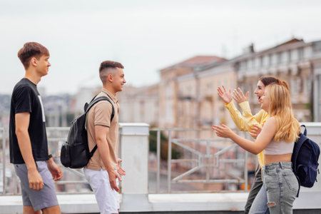 Four teenagers in casual clothes chat and laugh on the roof of the house. Girls and boys have fun together. Cute students with backpacks relax and walk.の写真素材