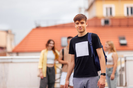 Close-up of a male portrait of a cute guy in a beige T-shirt with a black backpack. The teenager smiles and looks at the camera. Roofs of houses and other teens in the background.の写真素材