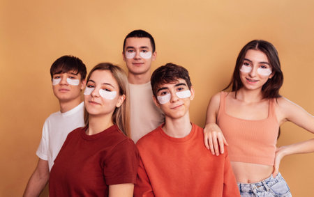 A group of smiling teenagers in casual clothes with moisturizing patches under their eyes. The five friends take care of the skin of the face. Happy girls and boys have fun together. Beige background.の写真素材