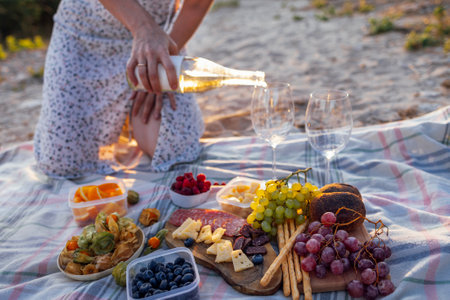 Close-up of a woman hand holding a bottle of wine and pouring white wine into glasses outdoors. Picnic on the beach. Delicious snacks, bread, fruits and berries on the bedspread.の写真素材