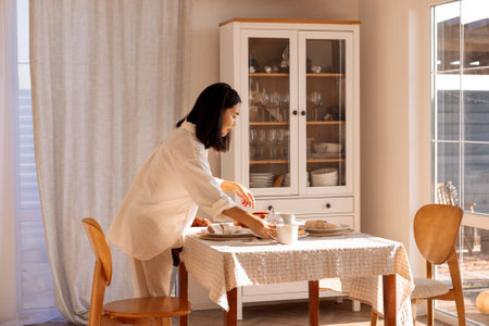 A young cute Asian woman sets the table in the dining room at home. A charming Korean girl sets out plates and cups for a family breakfast. Household chores and routine.の写真素材