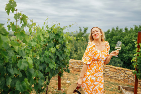 Cheerful blonde woman in bright orange dress with floral print holds glass of champagne high above her head in vineyard. Pretty girl in sunglasses laughs while looking at glass of sparkling wine.の写真素材
