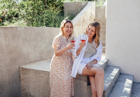 Young blonde women in casual clothes laughing and holding glass of champagne. Attractive girl sits on stone parapet and drinks delicious pink wine outdoors. Tourism, travel and friendship concept.の写真素材