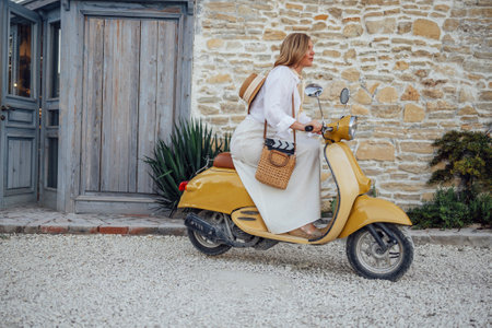 Beautiful female driver in white shirt and beige dress driving yellow motoroiler. Happy attractive woman in straw hat on the background of old stone wall. Smiling blonde on bike in town. Vintage styleの写真素材