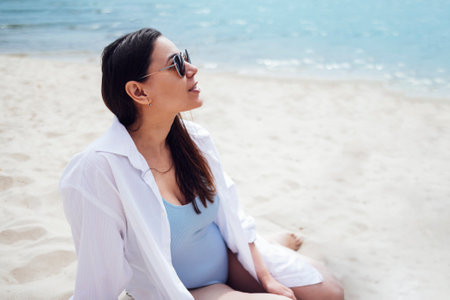 Attractive young woman in sunglasses, swimsuit and white shirt sits on a sandy beach. Charming brunette by the sea. Beautiful smiling girl in profile. Relax and vacation concept.の写真素材