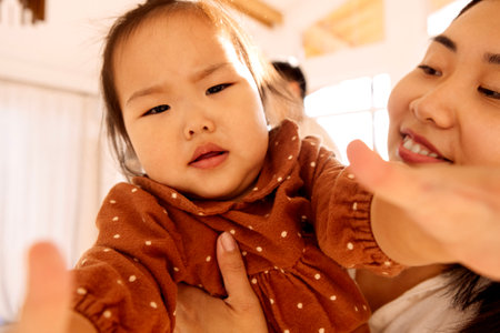 Cute Asian girl in a brown dress with her attractive mom. A smiling Korean woman holds her little daughter. Sweet toddler pulls her hands forward.の写真素材