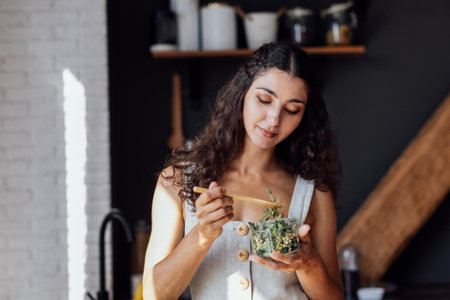 Attractive young woman is eating salad of sprouted greens in kitchen. Curly-haired brunette girl in linen sundress holds fork and transparent bowl with micro greenery. Healthy food concept.の写真素材