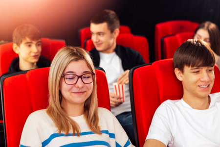 Nice young people in the cinema hall. A cute girl with glasses and popcorn and a guy are sitting in...
