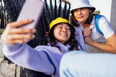 Two young charming multiethnic women take selfies and laugh outdoors. Smiling friends of different races in casual clothes and hats have fun and take pictures. An Asian girl is lying in a hammock.の写真素材