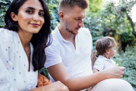 Cute family in light casual clothes in garden on terrace of house. Young couple and their outdoor sons on summer sunny day. Dad standing back and holding his toddler boy. Attractive woman is smiling.の写真素材