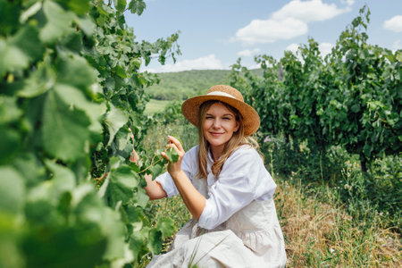 Young beautiful woman takes care of the vineyard. Caucasian pretty girl in beige sundress, white shirt and straw hat examines green bunches of grapes. Green rows with grapes in the background.の写真素材