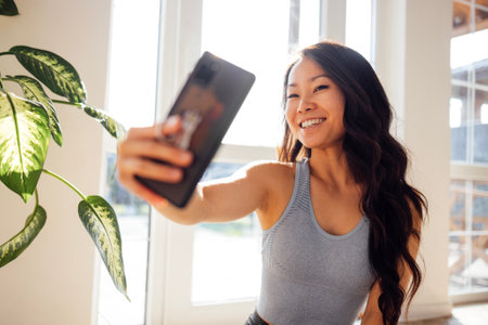 Charming Asian woman uses the phone at home in the living room. A pretty young female Korean holds a smartphone and communicates online. A cute girl writes a message. Stylish cozy sunny interior.の写真素材