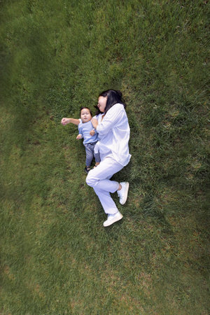 Asian smiling mom and sweet daughter are lying on a green lawn. A charming Korean woman and her cute kid are having fun on the grass. A young mother and her little baby in casual clothes in a meadow.の写真素材