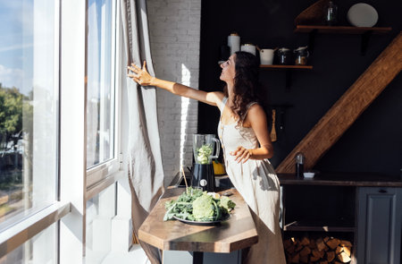 A beautiful curly-haired brunette in a linen sundress is cutting greens in the kitchen. A young attractive smiling vegetarian woman makes a green smoothie at home. Modern cozy sunny interior.の写真素材