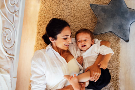 A young smiling woman and her little son are lying on the bedroom floor and having fun. An attractive mom and her sweet baby are laughing together. A happy mother tickles her cute toddler.の写真素材