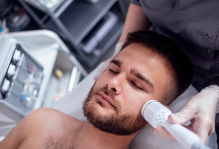 Closeup of face of beautiful relaxing man undergoing hardware cosmetology procedure in spa. Cream for ultrasonic facial massage is applied to face of young male using special nozzle in beauty salon.の写真素材