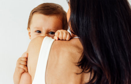 A young woman holds her beautiful baby. Charming mom with a sweet cute toddler on a white wall background. A mother and her little son are at home. Motherhood and caring concept.の写真素材