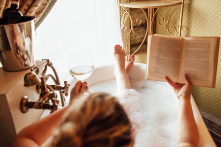 Close-up of a hand holding an elegant glass of champagne, reading the book in the bathroom. A young...