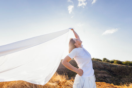 An attractive young woman smiles and holds an open white sheet outdoors. A charming girl enjoys the wind and freedom. A handsome lady is having fun in nature. A beautiful landscape.の写真素材