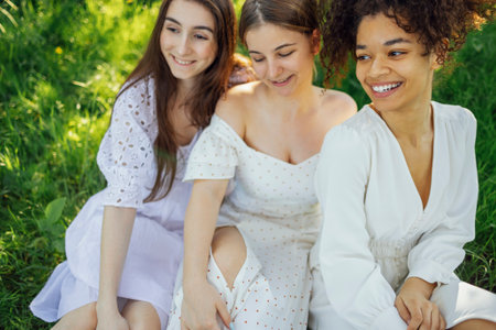 Three cute female teenagers of different nationalities in white elegant dresses sit on grass. Smiling girls have great time together on meadow. Caucasian and mixed race girls laugh and enjoy life.の写真素材