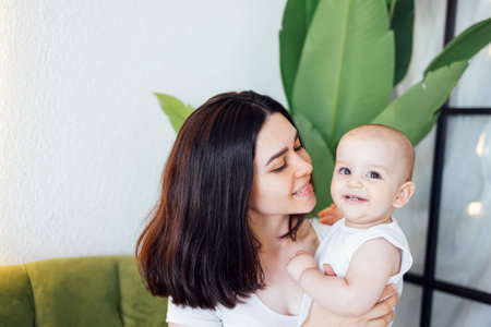 A beautiful mother hugs her cute baby indoors. A young woman holds her smiling child in her arms in...