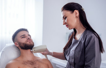 Young man receiving photo epilation while visiting beauty center on salon, professional female beautician doing laser hair removal procedure on clients face. Cosmetology conceptの写真素材