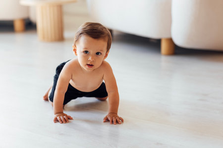 A cute baby is sitting on the white wooden floor in the living room. The little boy toddler is...