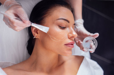 Beautiful brunette girl is doing a cosmetic procedure. The cosmetologist applies a rejuvenating white mask to the female client face with a brush. The concept of beauty and skin care. Close up.の写真素材