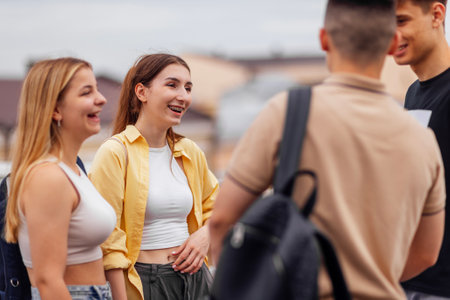 Four teenagers in casual clothes chat and laugh on the roof of the house. Girls and boys have fun together. Cute students with backpacks relax and walk.の写真素材