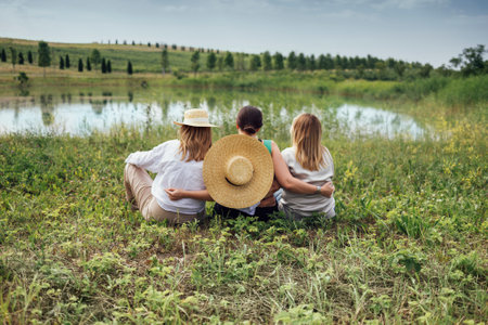 Three young women in casual clothes are sitting by shore of lake or pond and enjoying nature. Female friends hug and look at beautiful landscape with green hills and trees. Girls are relaxing togetherの写真素材