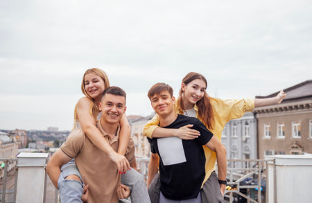 Cute girls in casual clothes ride on the backs of handsome guys on the roof of the house. Two young couples have fun. Happy teens enjoying vacation together. Smiling boys and girls on the street.の写真素材