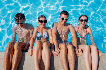 Cute teens friends in swimsuits and sunglasses are laughing in the pool. Smiling boys and girls having fun in the clear water. Joyful teenagers are relaxing together.の写真素材