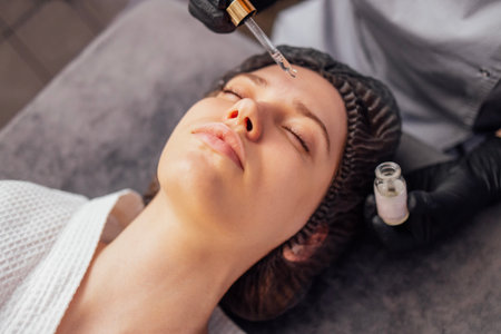 Beautician applies serum from pipette in black gloves to the face of client in spa salon. Close up...