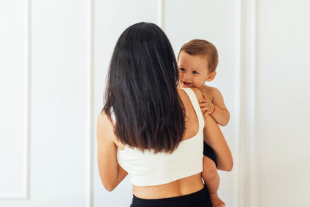 A young woman holds her beautiful baby. Charming mom with a sweet cute toddler on a white wall...
