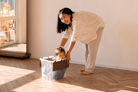 Young laughing Asian parent roll her daughter in a plastic box around the house. Korean mom and their cute little girl have fun together in the living room on a sunny day. A happy family.の写真素材