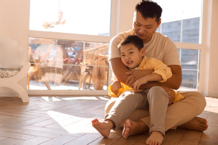A young happy Asian man hugs his little son. A Korean dad is playing with his child on the living room floor. A laughing father and his cute kid are having fun together at home.の写真素材