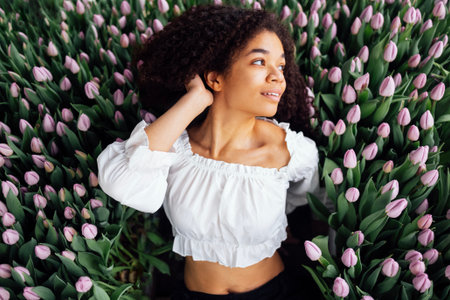 Close up of female portrait of cute charming African ethnicity girl among gently purple color tulip buds. Romantic young afro girl in white shirt touches fresh flowers with her face.の写真素材