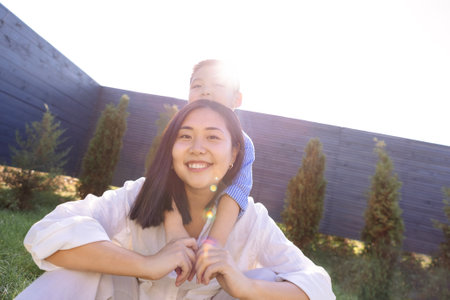 Asian laughing mom and son are hugging on a green lawn. A smiling young woman and her cute kid are having fun on the grass. A mother and her child in casual clothes in a meadow. View from above.の写真素材