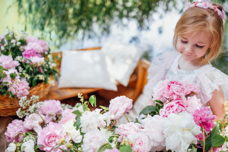 Portrait of a cute little girl in an elegant dress on a wooden boat among pink peonies on the pond or lake. A blond child plays outdoors in the park. A pretty kid is looking at flowers. Copy space.の写真素材