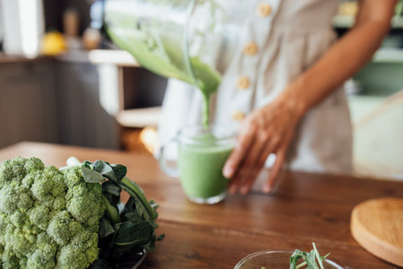 Close-up of broccoli on the kitchen table. A young woman pours a green smoothie from a blender into a glass cup. A vegetarian girl makes a healthy and delicious drink at home.の写真素材