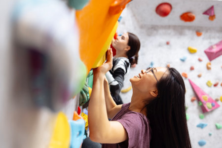 A group of multiracial women doing rock climbing in the gym. Attractive girls climb the bouldering wall. Professional sport female climbers train and laugh. Charming multiethnic friends are actively spending time.の写真素材