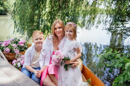 Beautiful young woman and her blonde cute children in elegant clothes pose for the camera. Family portrait near a pond, lake or river. A charming mother with two lovely children resting by the water.の写真素材