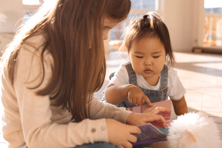 Cute children of different races play together. A Caucasian girl is reading a book to a sweet Korean baby. Little friends are having fun in the living room of the house.の写真素材