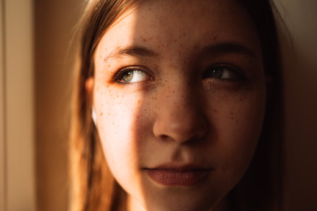 A close-up portrait of a cute teenager against the wall. A charming smiling girl with sunlight on her face. A pretty female teen in casual clothesの写真素材