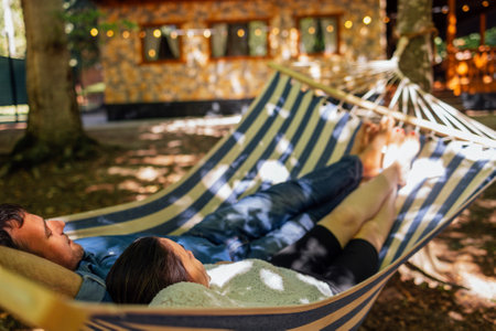 A young married couple is relaxing in the backyard of the house. An attractive husband and wife are lying in a hammock outside. A happy family spends time together and enjoys their holidaysの写真素材