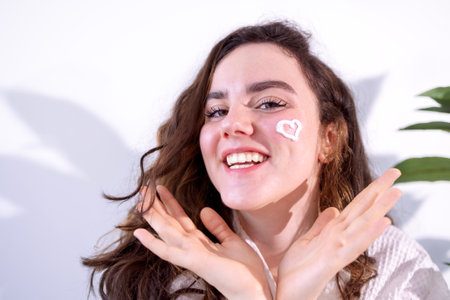 Close-up of a young attractive woman in a bathrobe laughing in a spa salon. A cream heart is painted on the face of a smiling red-haired girl. Beauty treatments and skin careの写真素材